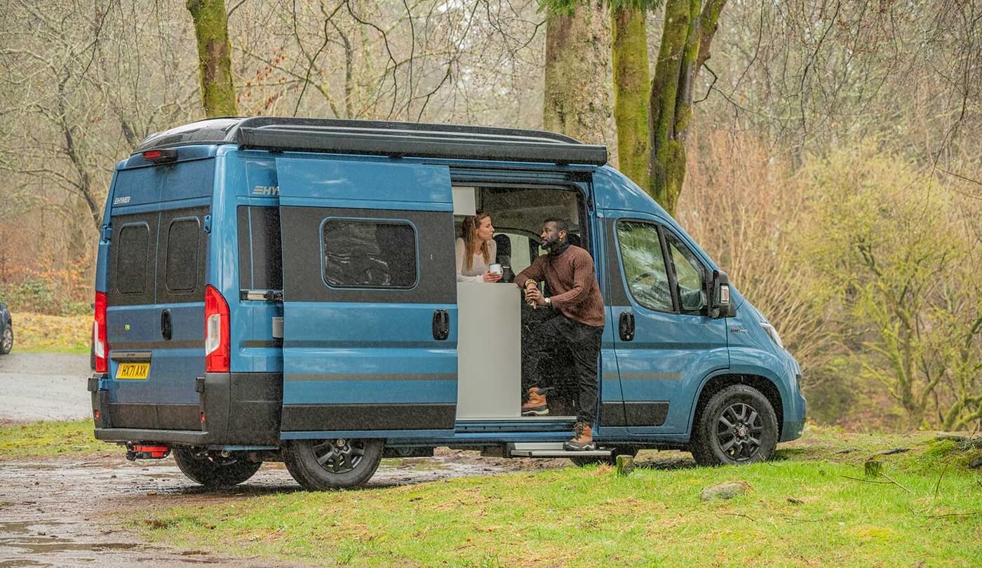 Deux personnes dans un fourgon bleu à la porte ouverte, près de la forêt.